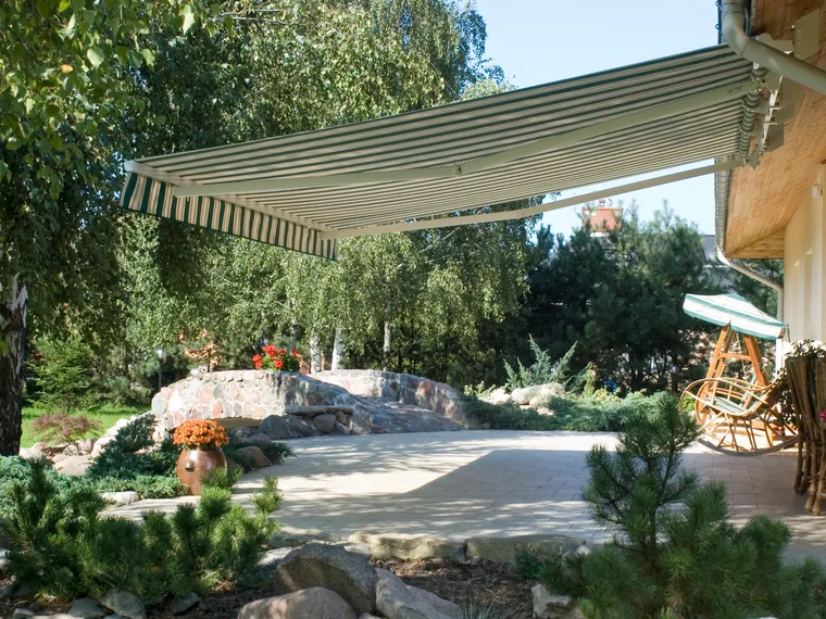 Zeleno-bijelo prugasta tenda nad terasom u vrtu s kamenjem, borovima i drvenom ljuljačkom