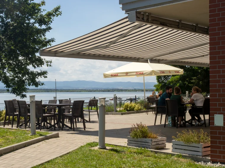 Tamno prugasta tenda nad terasom kafića uz jezero, ispod nje stolovi, suncobran i gosti