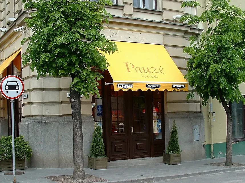 Žuta tenda Italia s volanom iznad ulaza u kafić na uglu ulice sa stablima